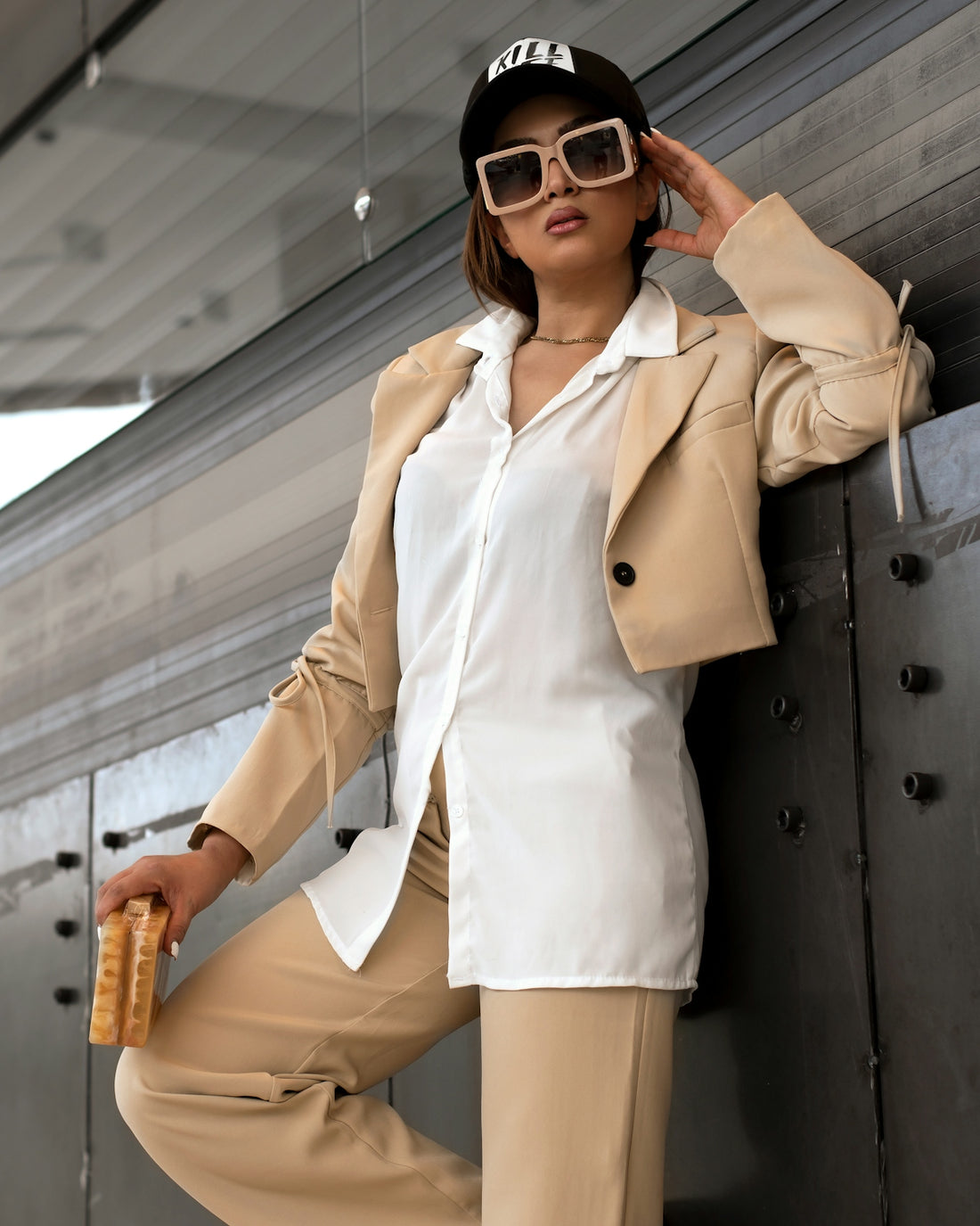 a woman leaning against a wall wearing a hat and sunglasses